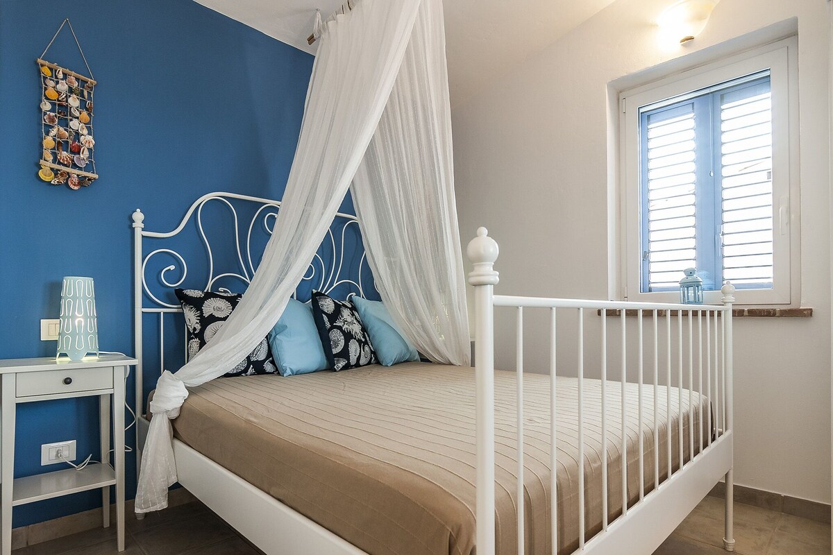Un dormitorio con estructura de cama metálica blanca, dosel, mesita de noche con lámpara y ventana con persianas.