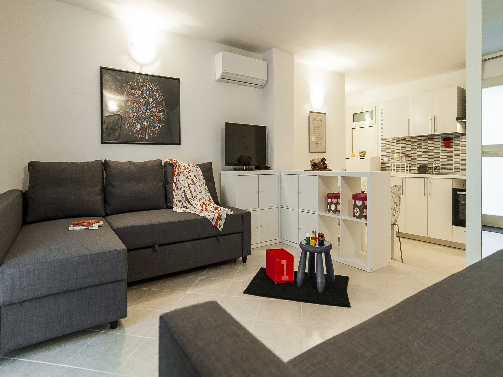 A modern living room with a grey sofa, a flat-screen TV, and a kitchen area with white cabinets and a tiled backsplash.