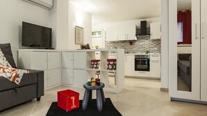 A modern living room with a grey sofa, a flat-screen TV, and a small table with toys. A red storage box is on the floor. The kitchen in the background has white cabinets and a tiled backsplash. - Casa Vacanza Cariddi Loft (Sperlonga)