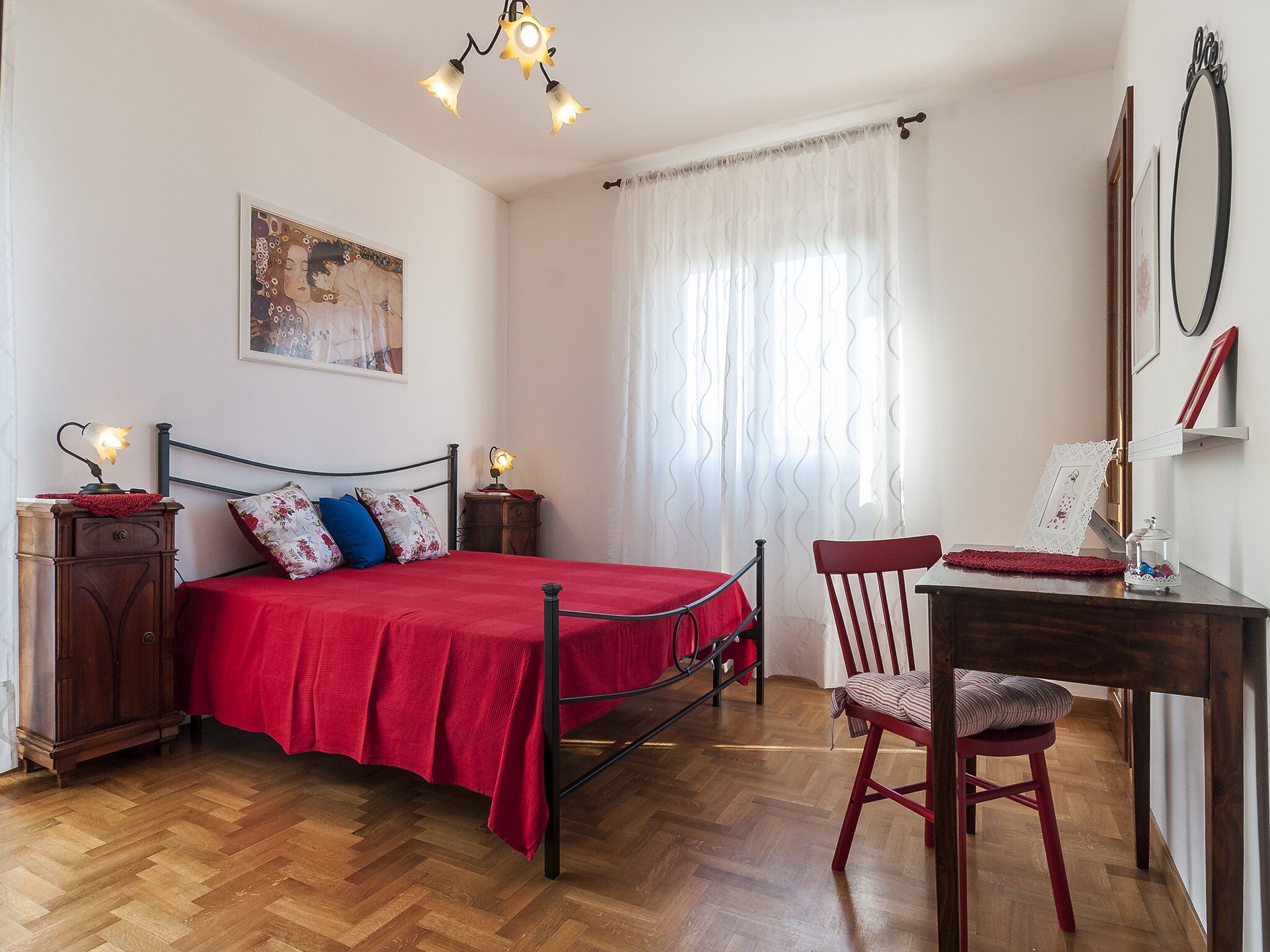 A bedroom with a wooden floor, a bed with a red bedspread, a nightstand with a lamp, a chair, a desk with a mirror, and a window with sheer curtains.