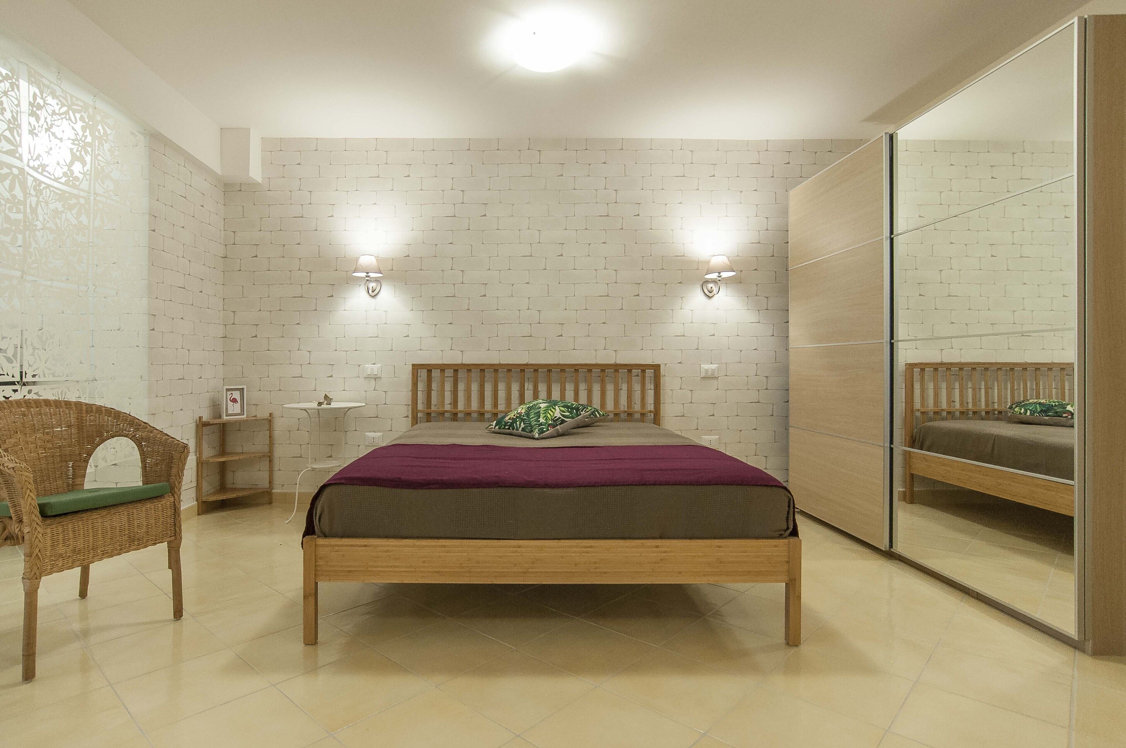 Une chambre avec un lit en bois, un oreiller vert, une couverture violette, une chaise en osier, une petite table et une étagère fixée au mur.