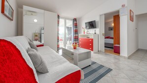 Una sala de estar moderna con un sofá rojo y blanco, una mesa de centro blanca y un mueble de almacenamiento rojo. Hay un televisor montado en la pared y una puerta que da a un balcón.