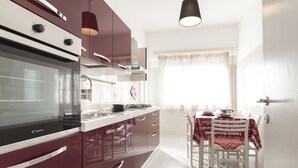 A modern kitchen with a built-in oven, a stove, and a sink. There is a dining table with chairs and a window with curtains. - Casa Vacanza Eolo (Sperlonga)