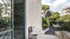 A building entrance with a wrought iron gate, a small outdoor seating area, and a pathway leading to a street with surrounding greenery and buildings. - Casa Vacanza Mare Verde (Sperlonga)
