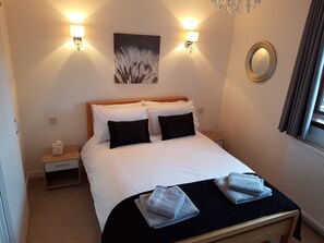 A neatly made bed with white and black bedding, two wall-mounted lamps, a painting, a mirror, and a small bedside table with a tissue box.