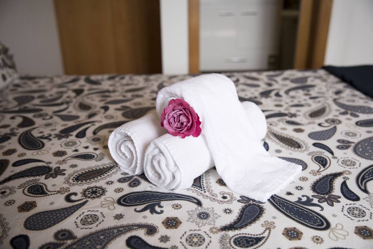 Un lit bien fait, recouvert d’une couvre-lit à motifs, avec une serviette blanche enroulée posée sur le dessus, ornée d’une fleur rose.