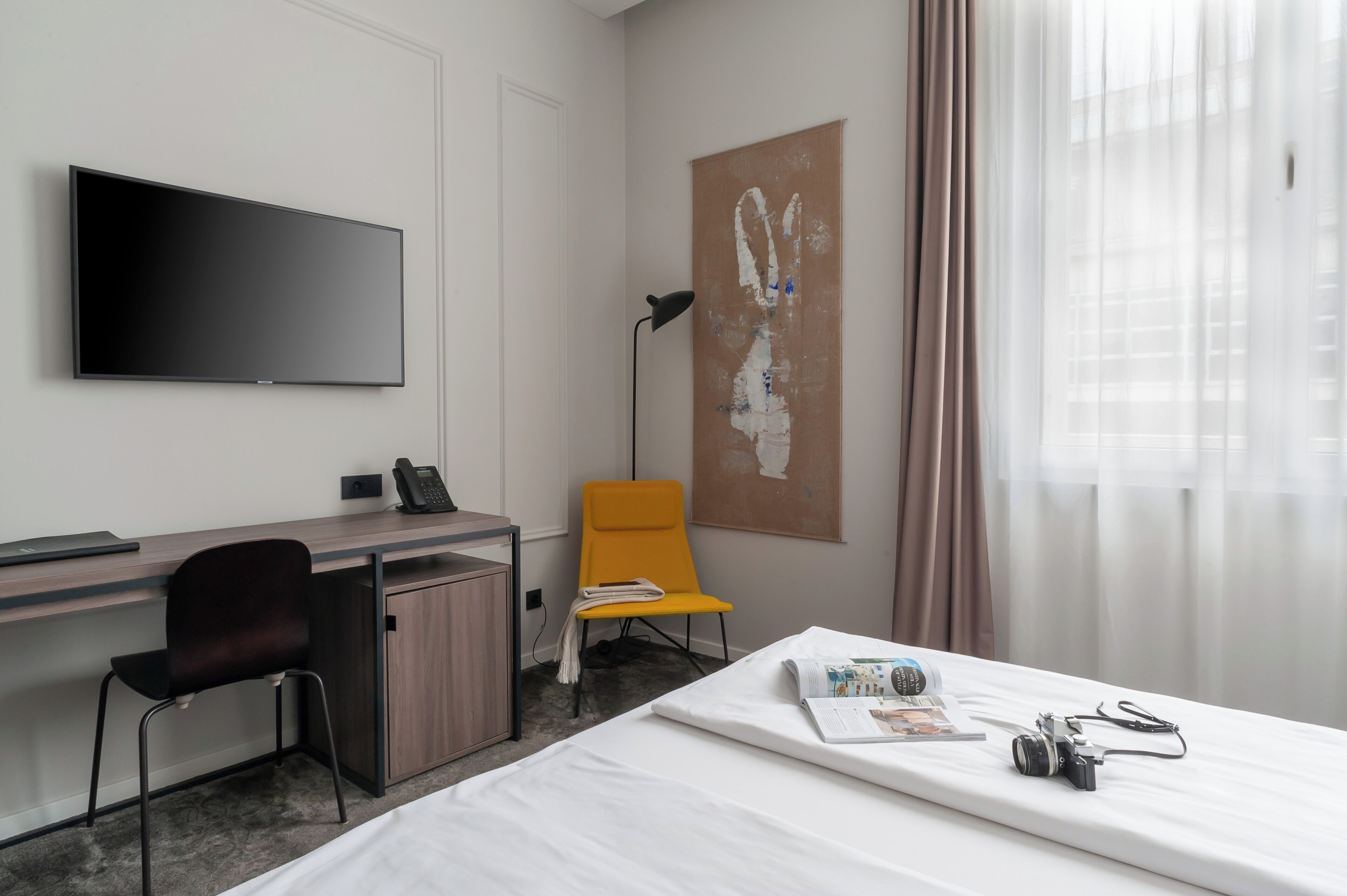 A hotel room with a bed, a desk with a chair, a flat-screen TV, a lamp, and a large abstract painting on the wall.