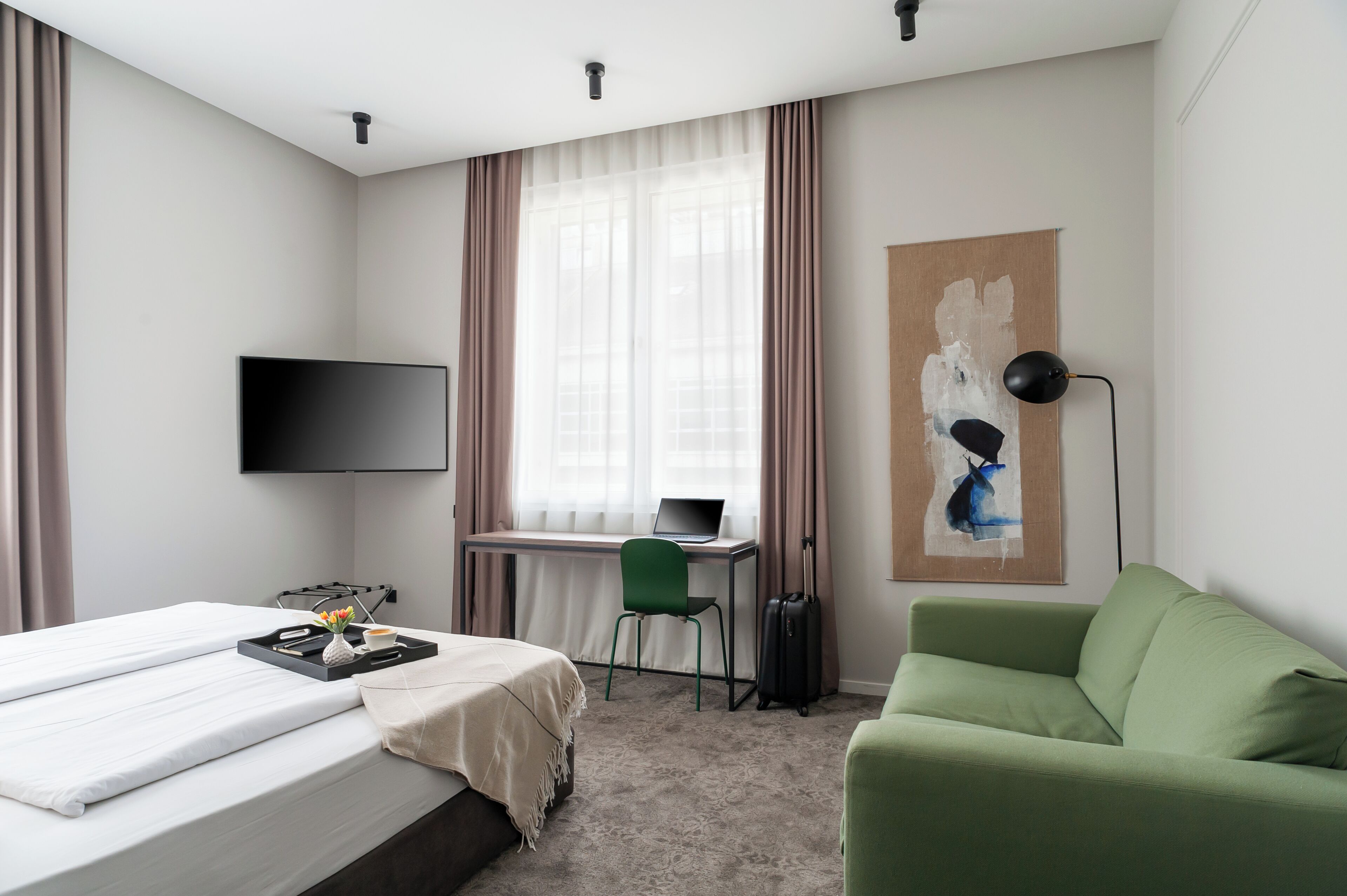 Habitación de hotel con una cama, un escritorio con una computadora portátil, una silla verde, un sofá verde, un televisor y un cuadro en la pared.