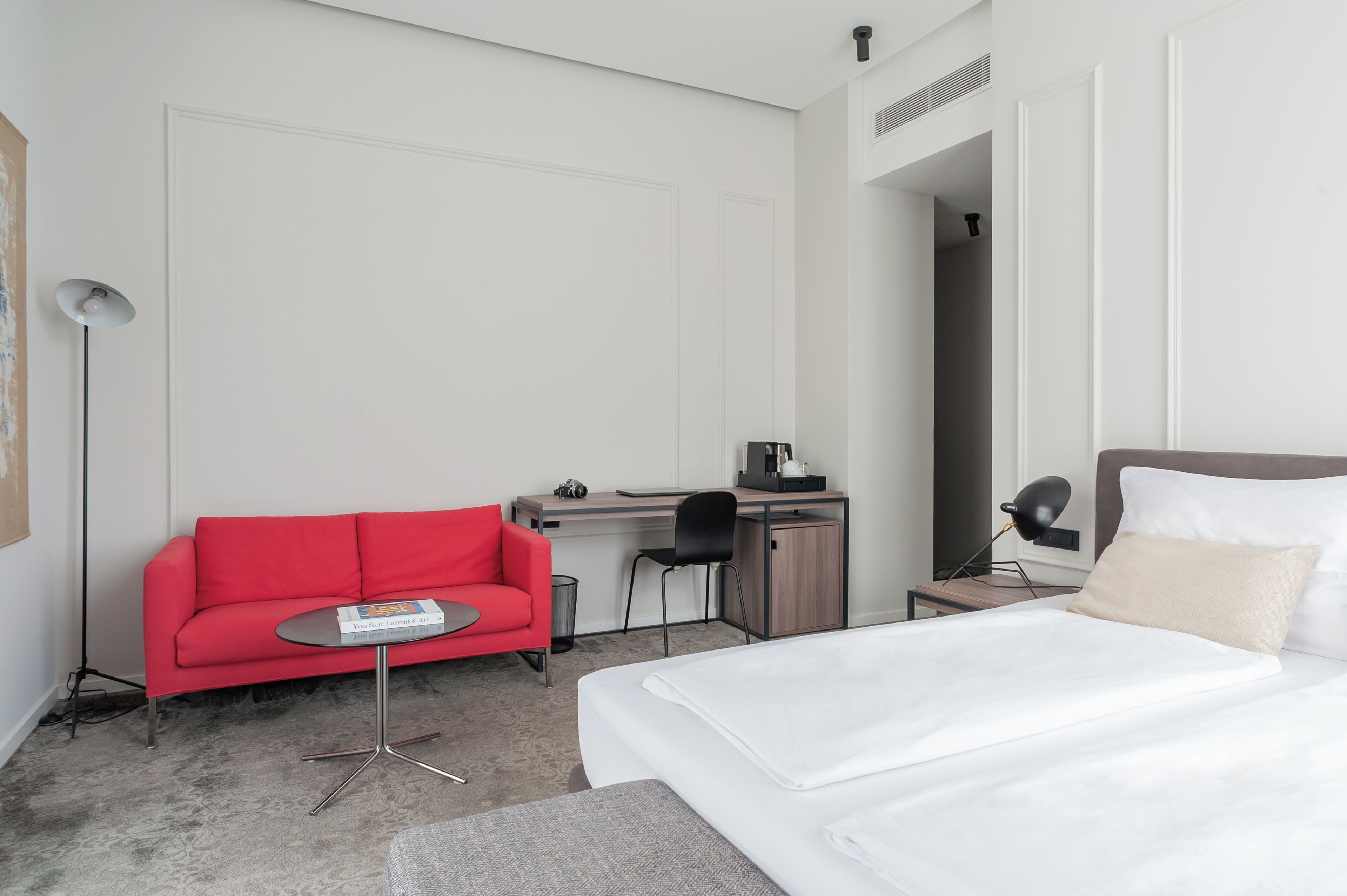 A hotel room with a bed, a red sofa, a desk with a chair, a lamp, and a small table.