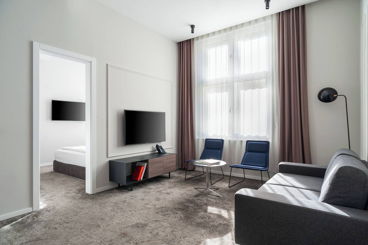 Habitación de hotel moderna con televisión de pantalla plana, sofá, dos sillones, una mesita y un amplio ventanal con cortinas.