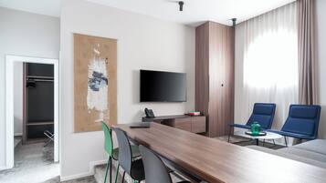 Habitación de hotel moderna con una mesa de comedor de madera, un televisor de pantalla plana, un sofá y un ventanal con cortinas.
