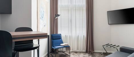 Et moderne hotelværelse med et træspisebord, en blå stol, en grå sofa, et sort tv ophængt på væggen og et vindue med lette gardiner.