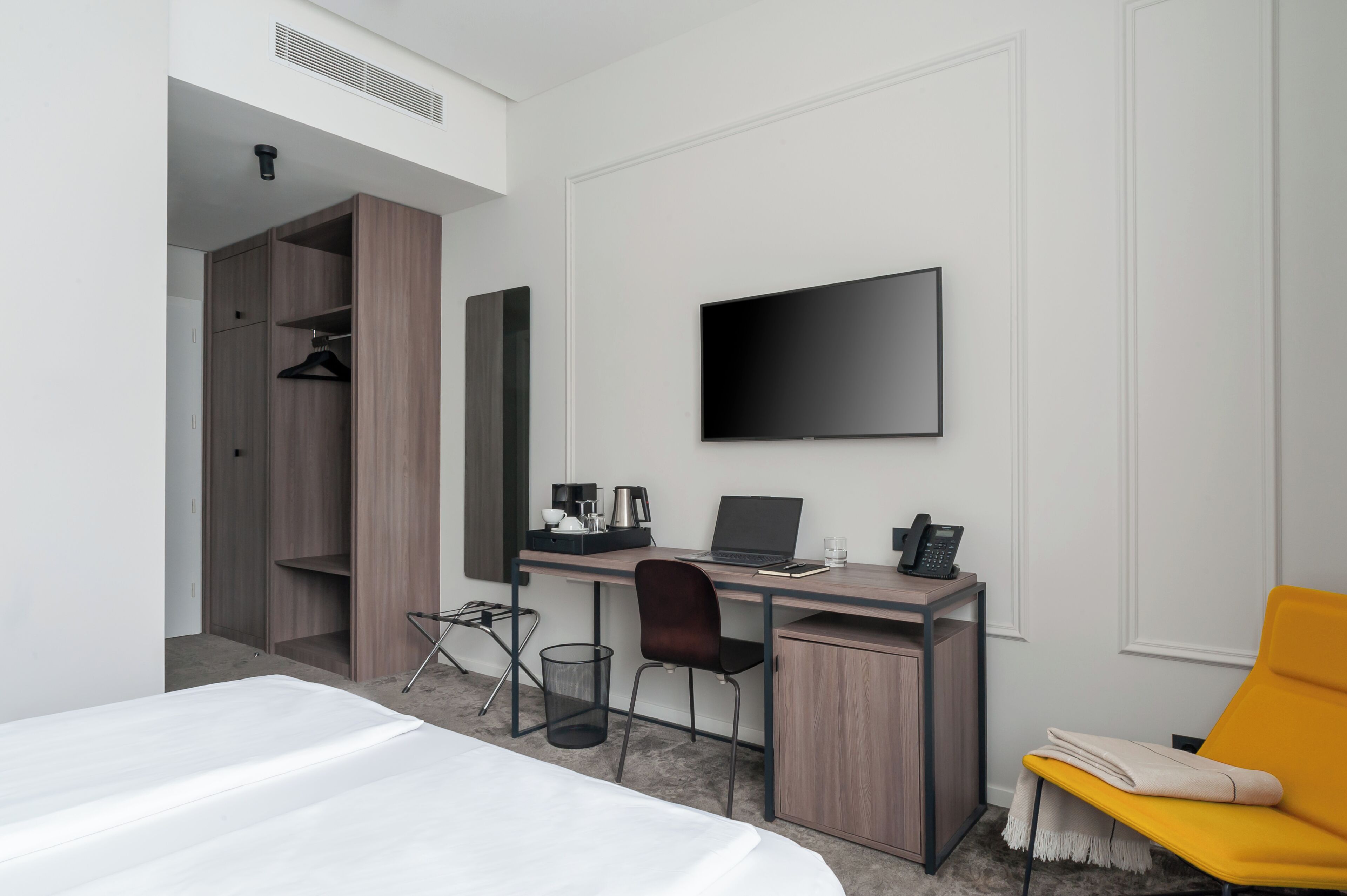 Quarto de hotel moderno com cama, escrivaninha com laptop, TV de tela plana, poltrona, armário e uma pequena lixeira.