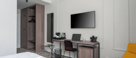 A modern hotel room with a bed, a desk with a laptop, a flat-screen TV, a chair, a wardrobe, and a small trash bin.