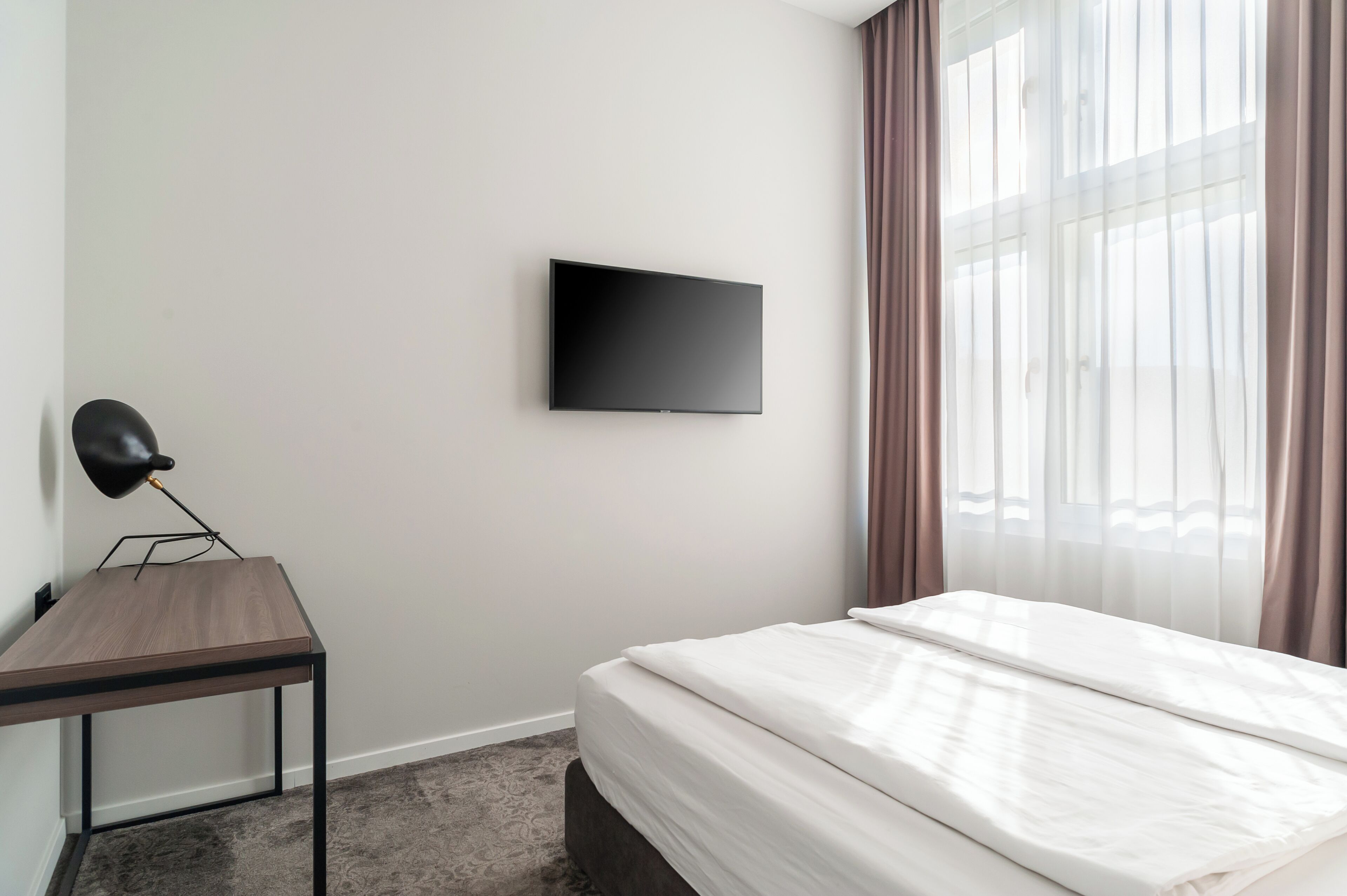 Une chambre d’hôtel équipée d’un lit, d’un bureau, d’un téléviseur à écran plat fixé au mur et d’une fenêtre avec des rideaux.