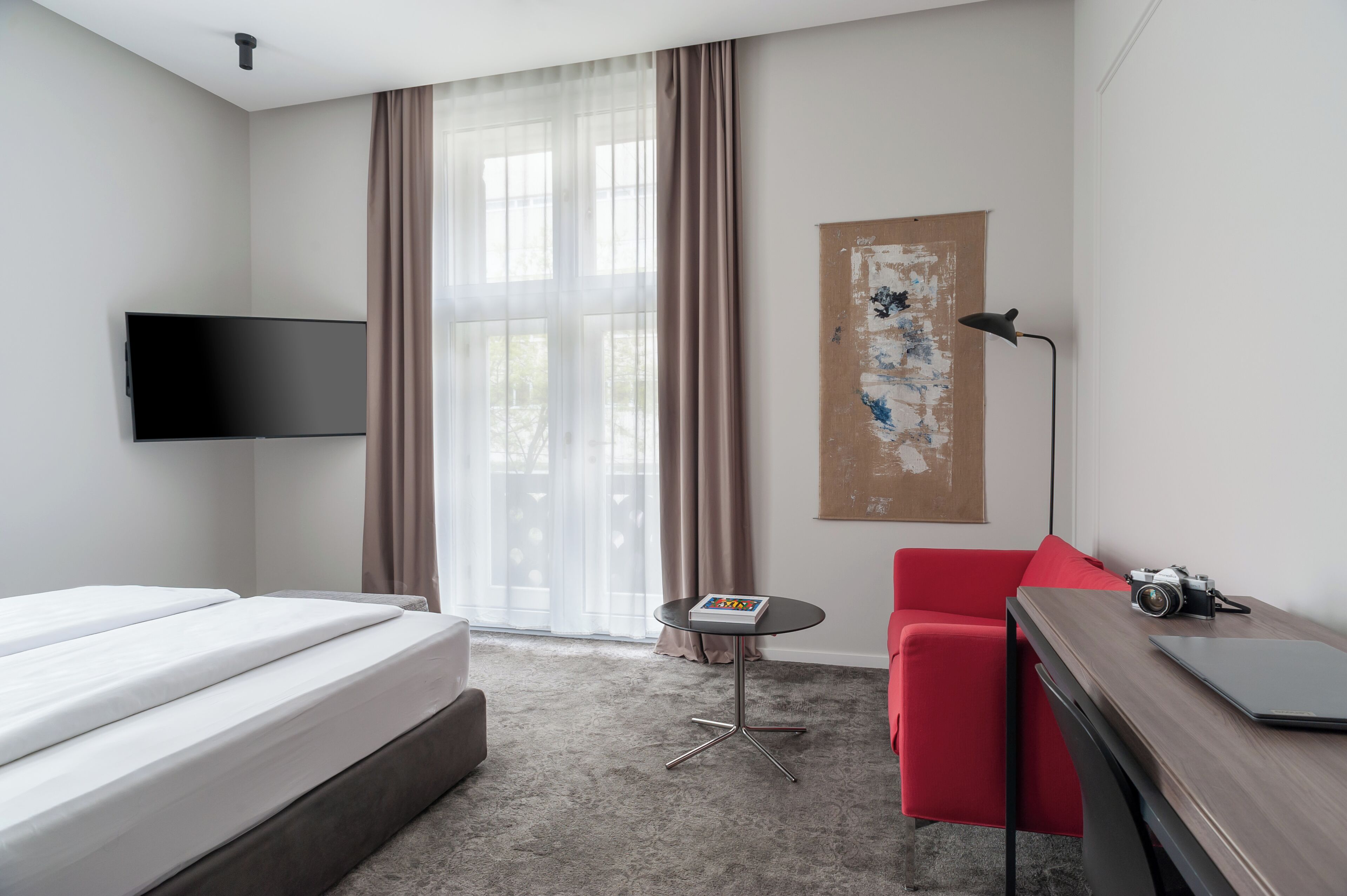 Una habitación de hotel con una cama, una silla roja, una mesita con un libro, un escritorio con una cámara y un cuadro en la pared.