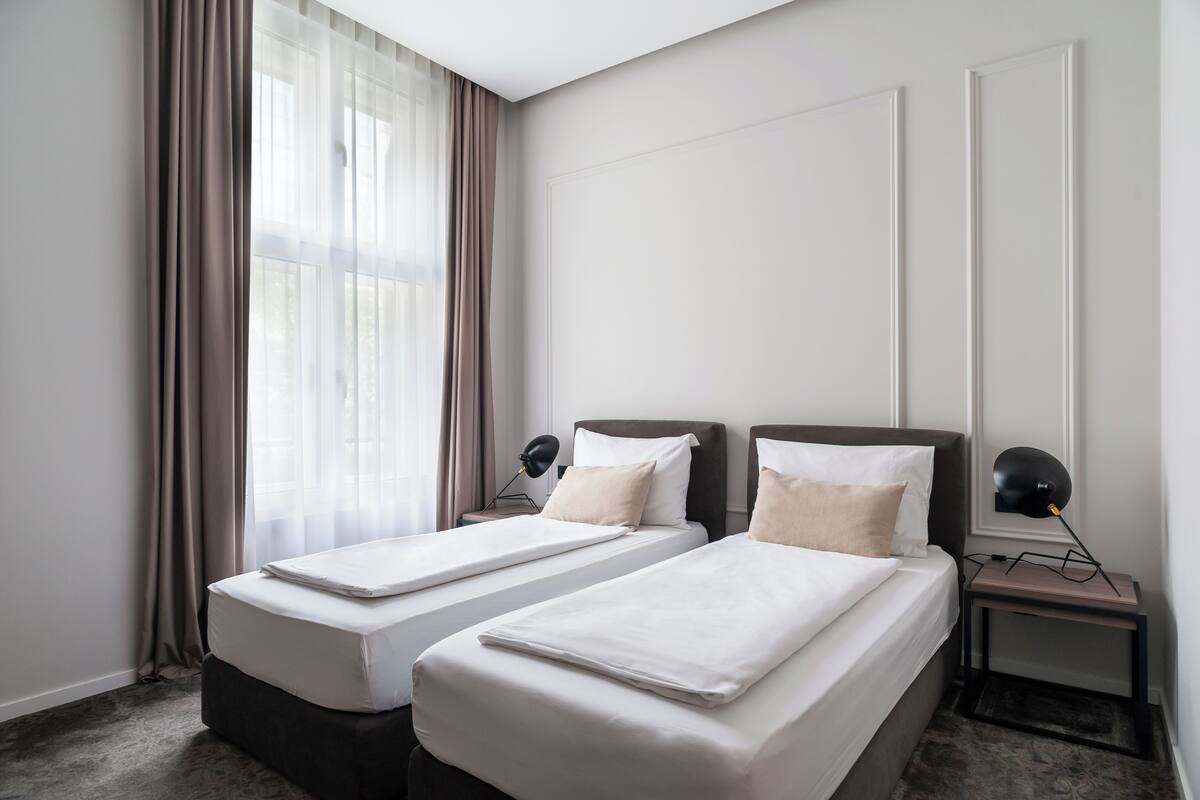 Una habitación de hotel con dos camas individuales, una mesita de noche con lámpara y un ventanal con cortinas.