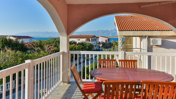 A balcony with a round wooden table and chairs, a white railing, and a view of the sea and mountains. - Apartement Delac (Vir)