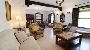 Un salon comprenant un canapé, des fauteuils, une table basse, une table à manger avec des chaises, un lustre et une grande fenêtre.
