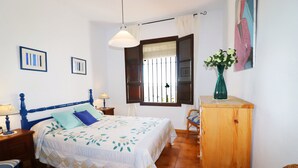 Une chambre à coucher comprenant un lit, une tête de lit en bois, des tables de chevet, une commode, une chaise, un meuble en bois, une fenêtre avec des rideaux et des œuvres d’art murales.