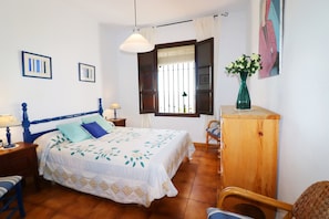 A bedroom with a bed, wooden headboard, bedside tables, a nightstand, a chair, a wooden cabinet, a window with curtains, and wall art. - 1146 Villa Hermoso (Nerja)
