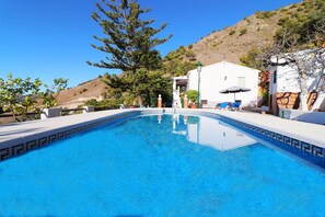 A swimming pool with a tiled edge, surrounded by a white building with a door and windows, and a tree in the background.