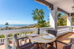 Una terrazza con tavolo apparecchiato per due, vista sulla costa, palme e una città in lontananza.