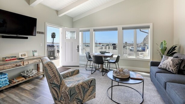 A spacious living area with a high ceiling, a flat-screen TV mounted on the wall, a dining table with chairs, a sofa, and a view of the city through large windows.