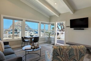 A spacious living room with large windows, a flat-screen TV, and a dining area with a table and chairs. - Morro Rock views, rooftop dining ~ Salty Sister 1Bdrm condo (Morro Bay)