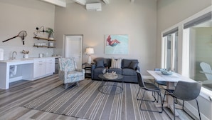 Un salon moderne avec un canapé gris, une table basse et une table à manger avec des chaises. On y trouve également un coin cuisine avec des meubles blancs et un plan de travail. Une grande fenêtre laisse entrer la lumière naturelle, et un tableau est accroché au mur.