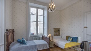 Een slaapkamer met twee bedden, een radiator, een raam, een kroonluchter en een waterkoker op een tafel.