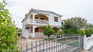Una casa de dos pisos con balcón, rodeada por una cerca de metal y follaje.