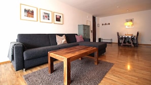 Ein modernes Wohnzimmer mit einem grauen Sofa, einem Holztisch und einem grauen Teppich. An der Wand hängen gerahmte Bilder, und im Hintergrund steht ein kleiner Tisch mit einer Lampe.