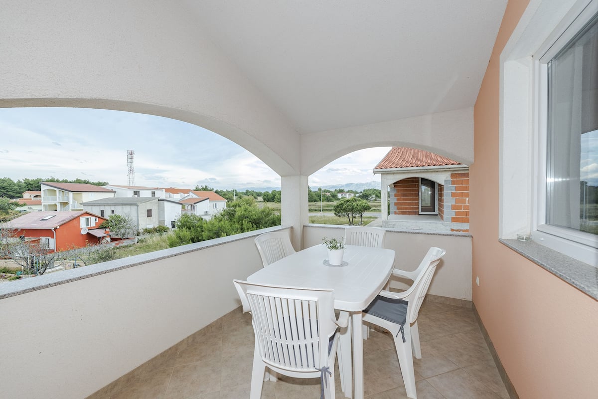 A balcony with a white table and chairs, a potted plant, and a view of neighboring houses and greenery.
