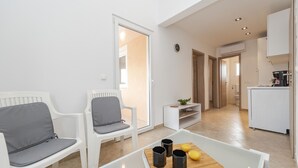 A modern living room with white furniture, a coffee table with fruits and cups, and a view to the kitchen and outside. - Apartments Simic (Vir)