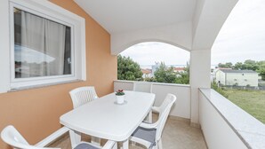 A balcony with a white table and chairs, a window with curtains, and a view of greenery and buildings. - Apartments Simic (Vir)