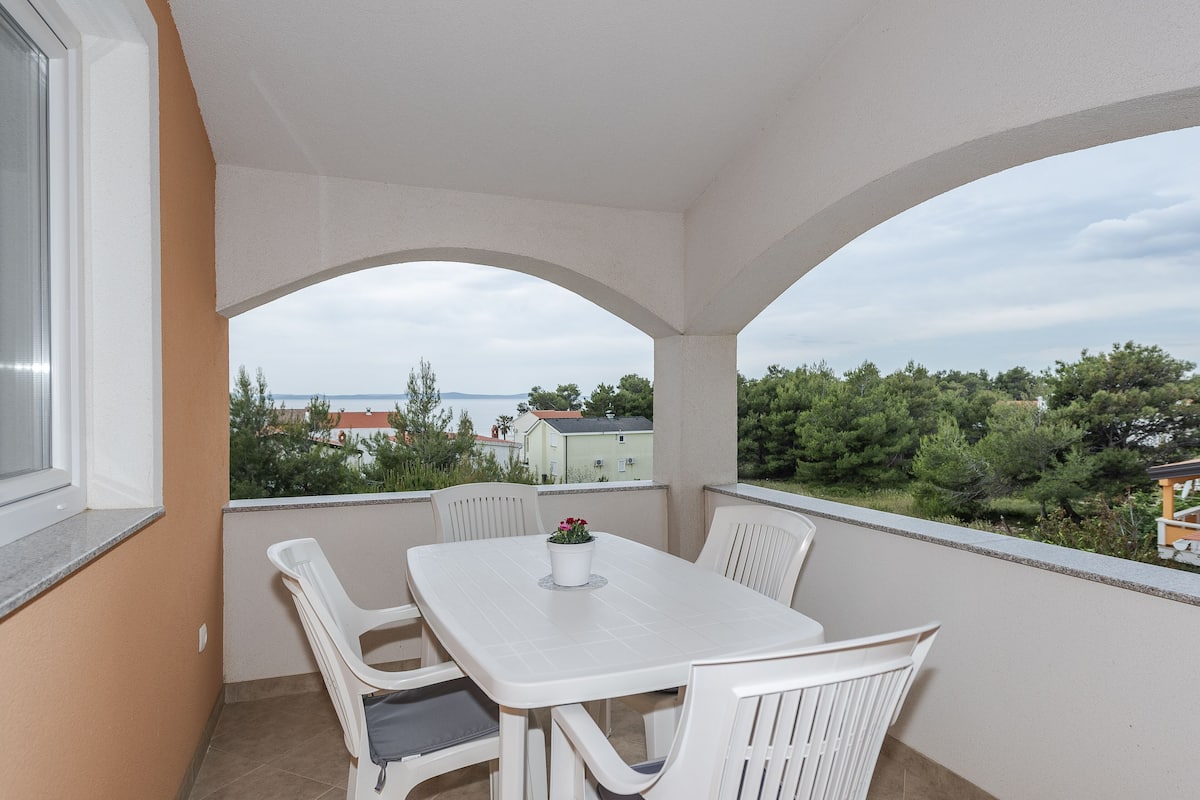 A balcony with a white table and chairs, a potted plant, and a view of the surrounding area.