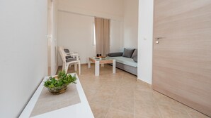 A modern living room with a sofa, a coffee table, and a chair. There is a door on the right and a window with curtains on the left. - Apartments Simic (Vir)