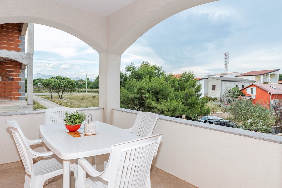 A balcony with a white table and chairs, a potted plant, and a view of a residential area with trees and houses.