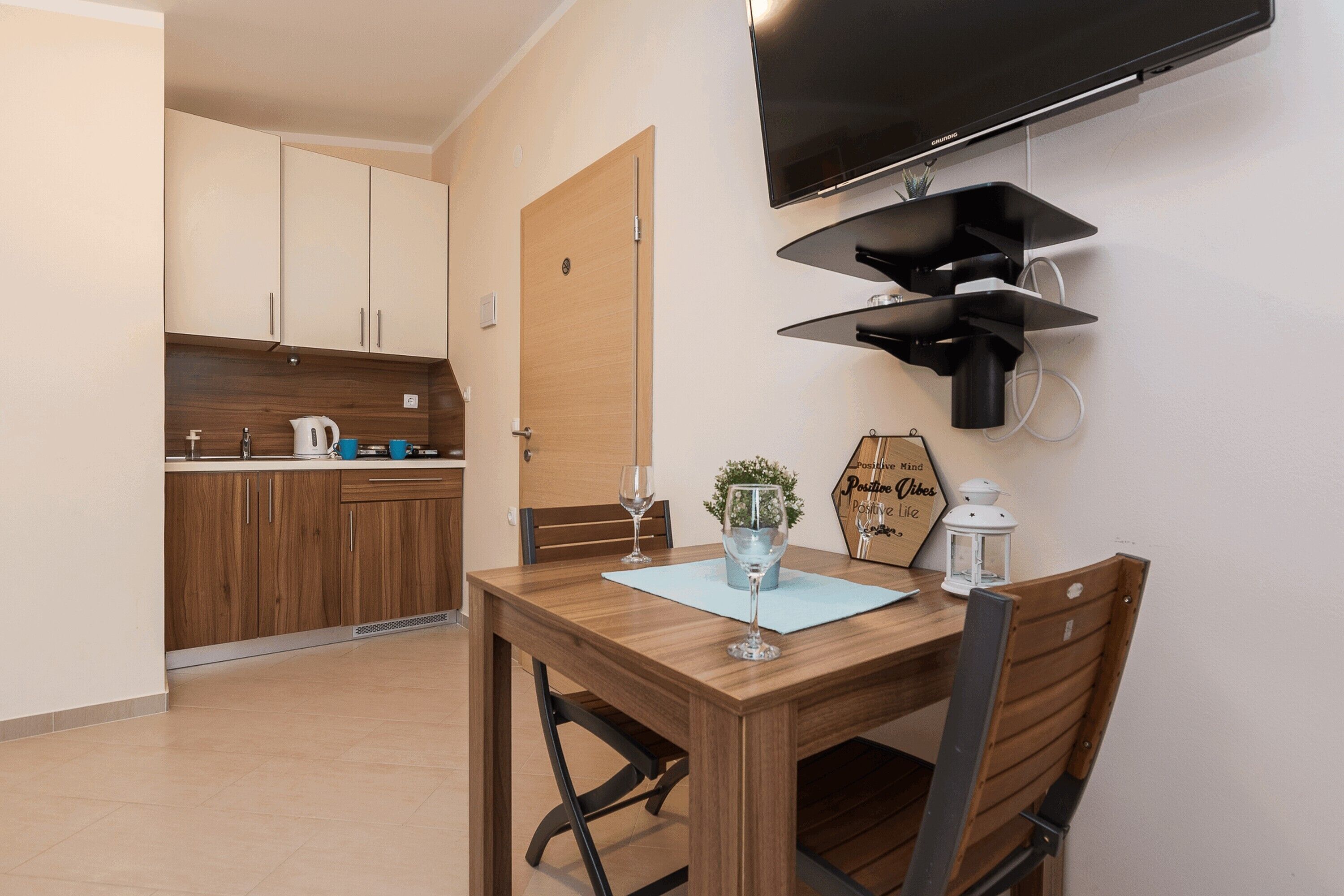 A compact kitchen with wooden cabinets, a dining table with chairs, a flat-screen TV mounted on the wall, and a small potted plant on the table.