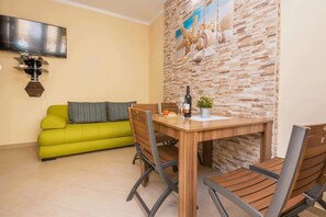 A dining area with a wooden table, chairs, and a green sofa. A television is mounted on the wall above the sofa. - Apartments Bobeta (Vir)