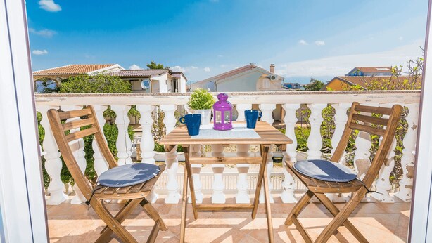 Ein Holztisch mit zwei Stühlen auf einem Balkon mit weißem Geländer, einem violetten Laternen und blauen Tassen.