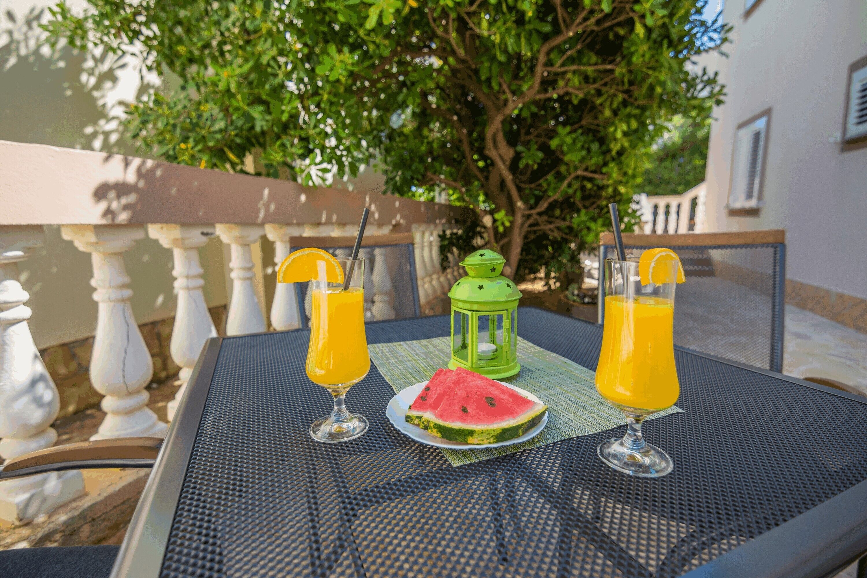 A table with two glasses of orange juice, a slice of watermelon, and a green lantern.