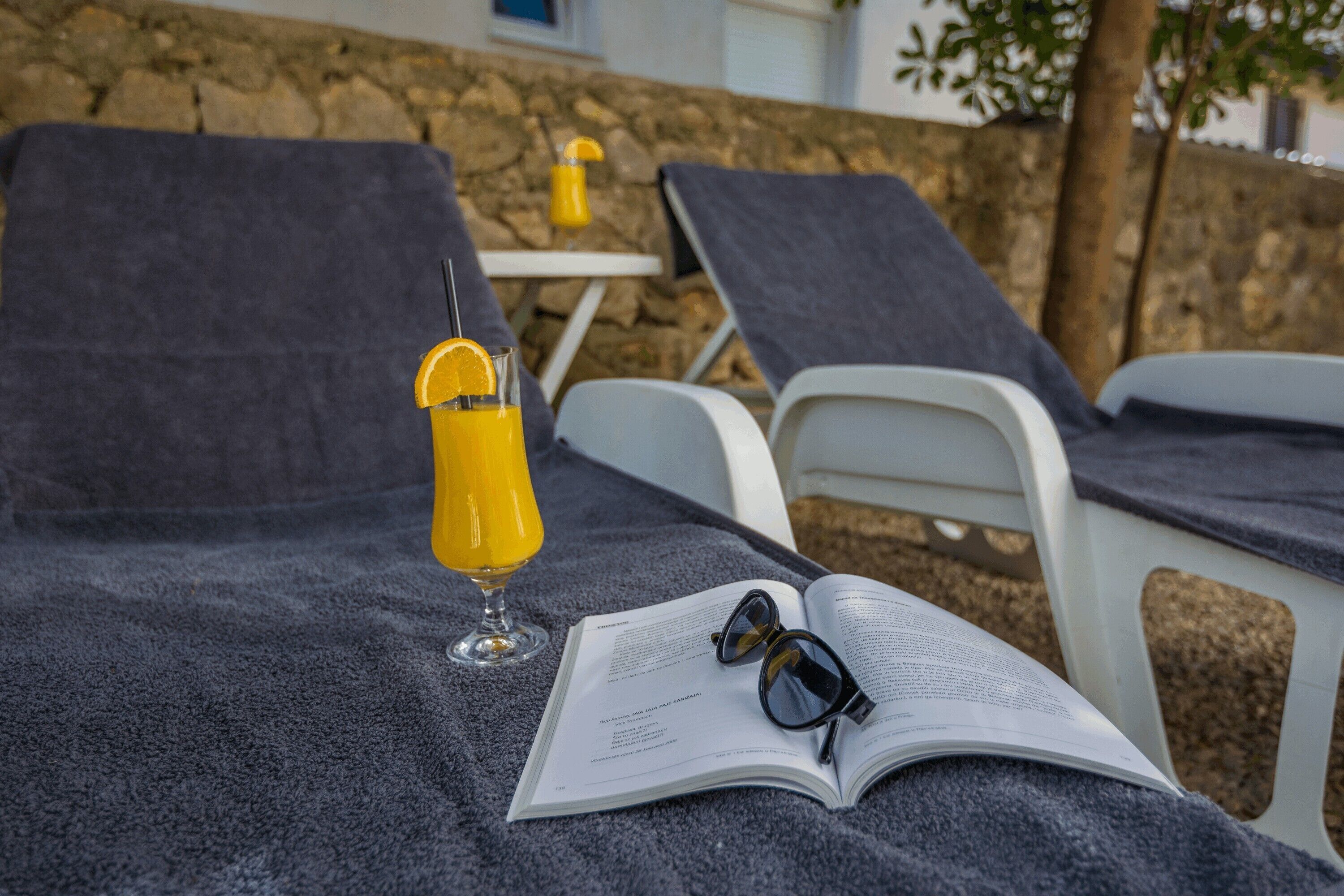 Una silla de descanso con un libro y gafas de sol, un vaso de jugo de naranja y una mesita con una lámpara.
