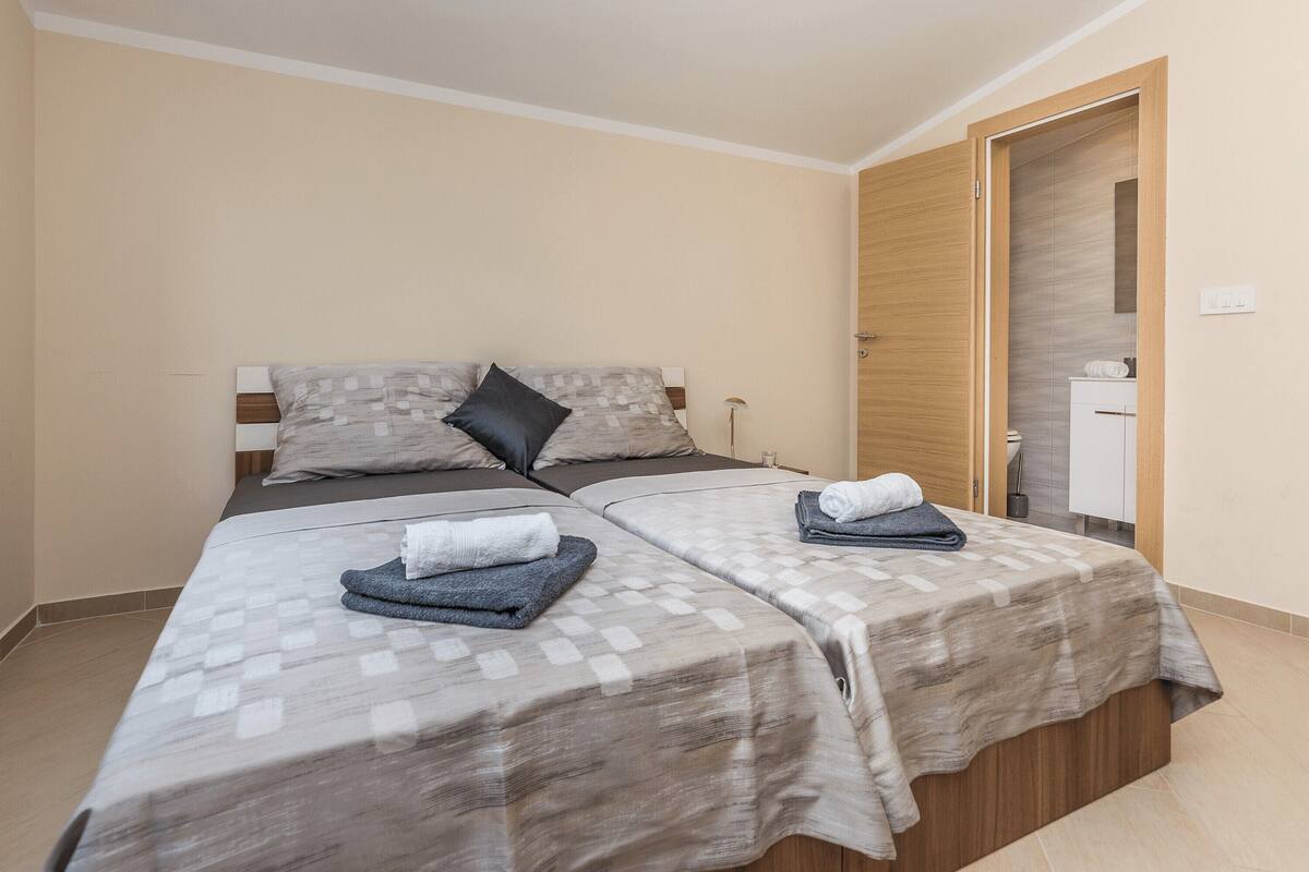 A double bed with a checkered bedspread and two pillows, a bedside table with a lamp, and a wooden door leading to a bathroom.