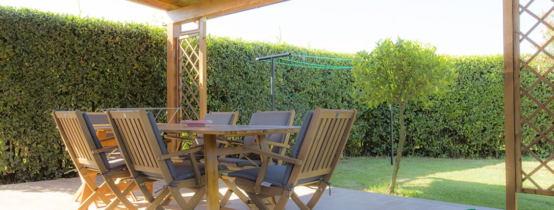 An outdoor patio with a wooden table and chairs, surrounded by a hedge.