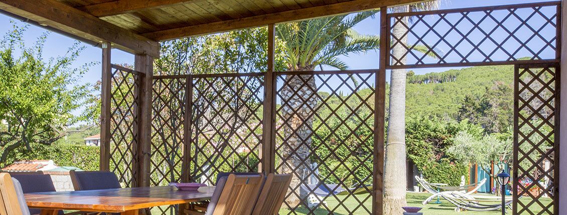 A covered outdoor seating area with a table and chairs, surrounded by a lattice fence, overlooking a lush garden and hills.