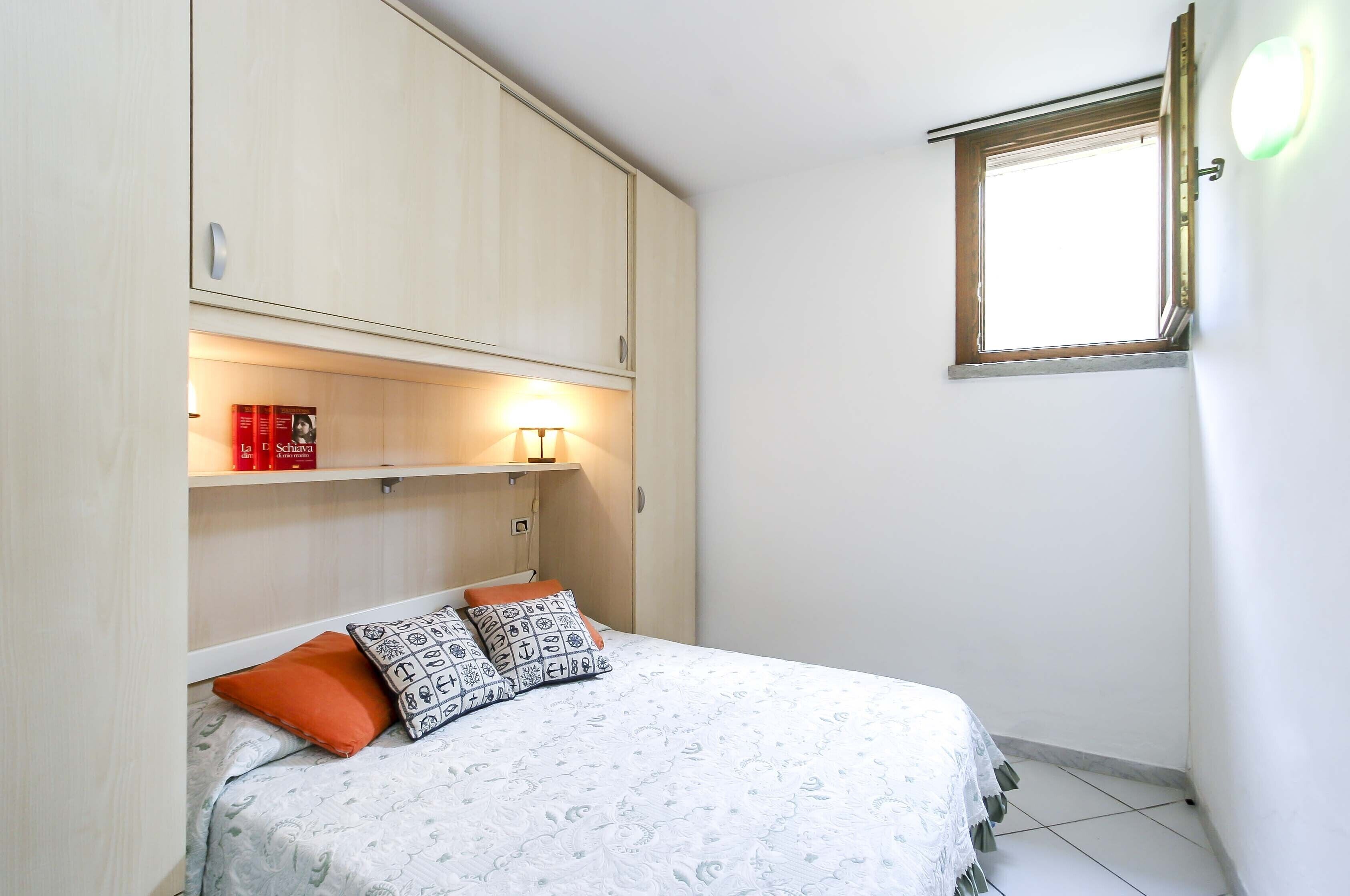 A bedroom with a bed, pillows, a shelf with books, a window, and a wall-mounted light.