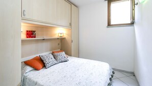 Une chambre à coucher avec un lit, des oreillers, une étagère remplie de livres, une fenêtre et un luminaire fixé au mur.