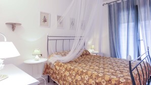 A bedroom with a bed, a bedside table, a lamp, a wall-mounted shelf, and a window with curtains. - Il Porto Bilocale 5 (Porto Azzurro)